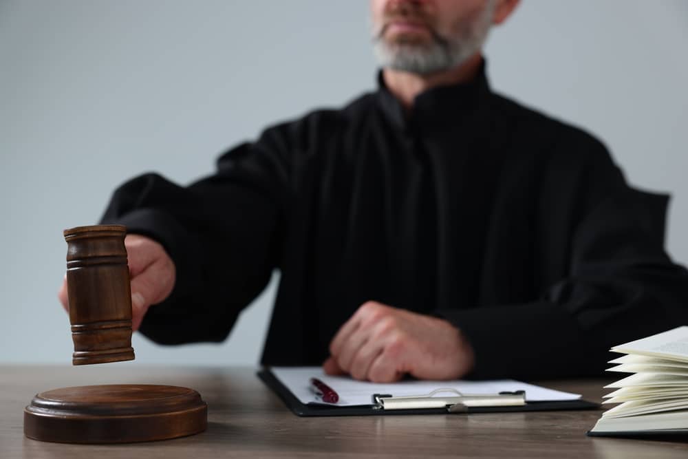 Judge with gavel and papers sitting at wooden table against light grey background, closeup